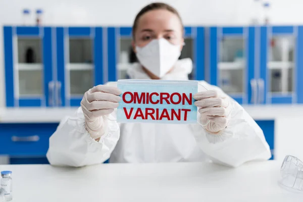 Blurred scientist in hazmat suit holding protective mask with omicron variant lettering — Fotografia de Stock