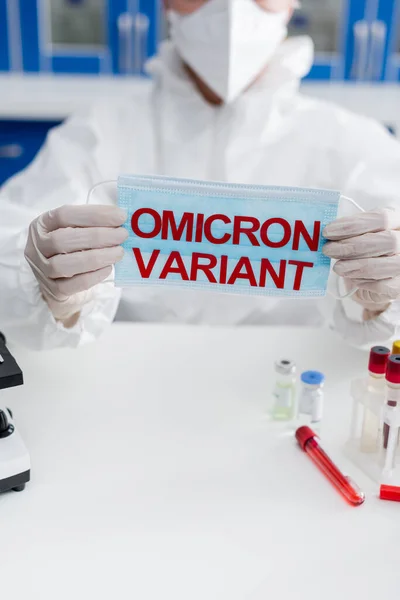 Partial view of blurred doctor in latex gloves holding medical mask with omicron variant lettering near test tubes and vaccine vials — стоковое фото