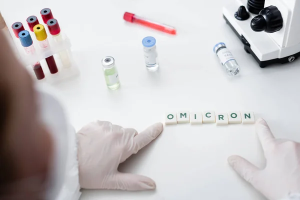 Partial view of blurred scientist near cubes with omicron lettering, vaccine vials and test tubes in lab — Fotografia de Stock