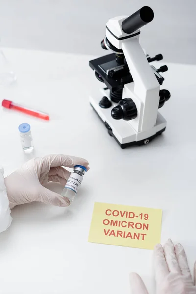 Vue recadrée du scientifique tenant le vaccin contre le coronavirus près du microscope et carte avec lettrage covid-19 omicron variante — Photo de stock