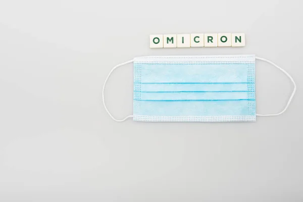 Top view of medical mask near white cubes with omicron lettering on grey background — Stock Photo