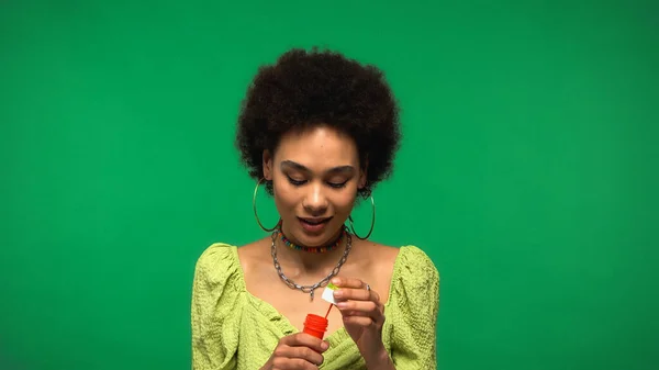Encaracolado afro-americano mulher segurando garrafa com bolhas de sabão isolado em verde — Fotografia de Stock
