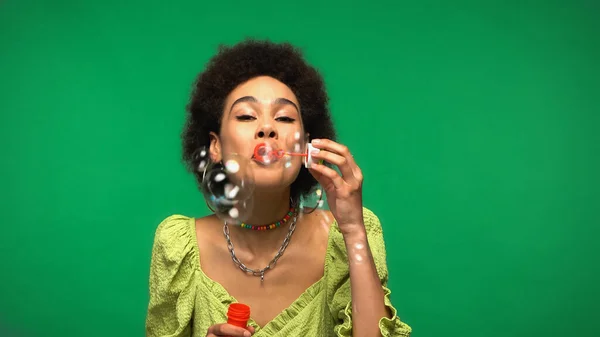 Curly african american woman blowing soap bubbles isolated on green — Stock Photo