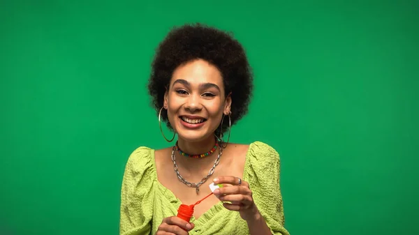 Happy african american woman holding bottle with soap bubbles and bubble wand isolated on green — Stock Photo
