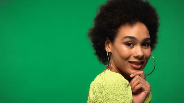 Alegre africana americana mujer en aros pendientes mirando cámara aislada en verde - foto de stock