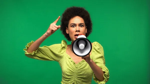 Mujer afroamericana sosteniendo megáfono y señalando con el dedo mientras advierte aislado en verde - foto de stock
