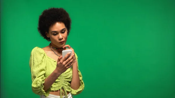 Задумчивая африканская американка, использующая смартфон, изолированный на зеленом — стоковое фото