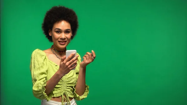 Happy african american woman using smartphone isolated on green — стоковое фото