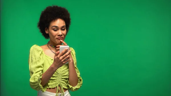 Задумлива афроамериканська жінка використовує смартфон ізольовано на зеленому — стокове фото