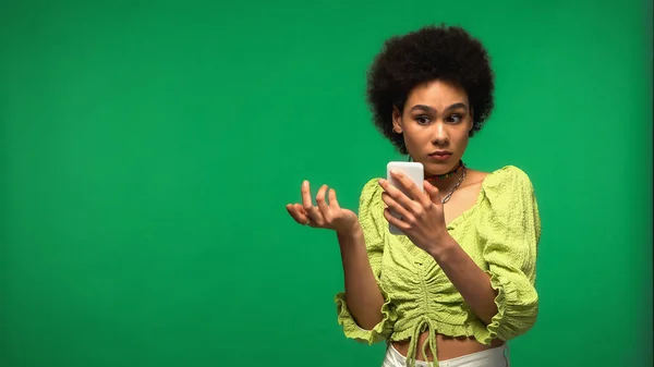 Dissatisfied african american woman using smartphone isolated on green — Stock Photo