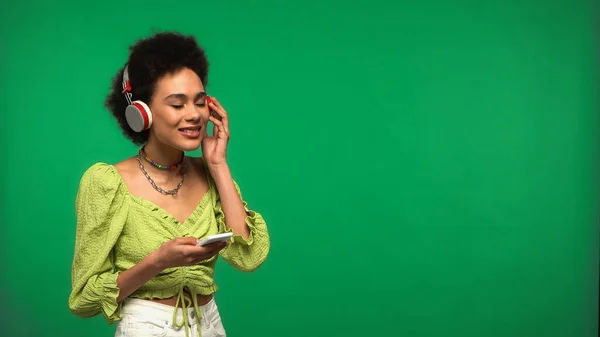 Pleased african american woman in wireless headphones holding smartphone isolated on green — Foto stock