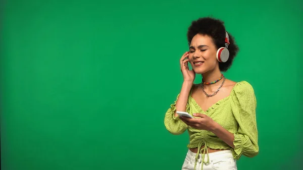 Happy african american woman in wireless headphones holding smartphone isolated on green — Stock Photo