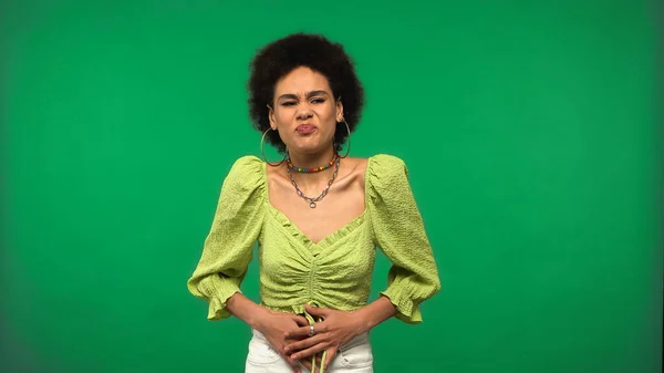 Displeased african american woman in hoop earrings having stomach ache isolated on green — стоковое фото