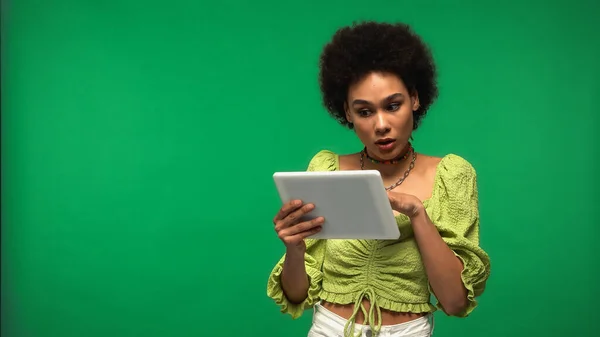 Surprised african american woman in blouse using digital tablet isolated on green — стокове фото