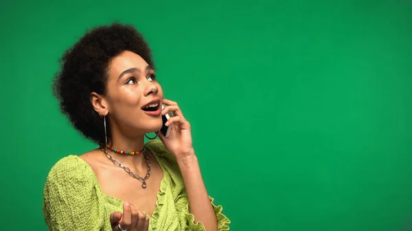Mujer afroamericana sorprendida en blusa hablando en teléfono inteligente aislado en verde - foto de stock