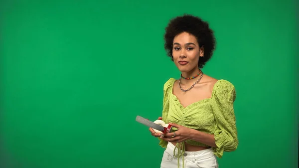 Curly african american woman in blouse holding wireless headphones isolated on green — Stockfoto