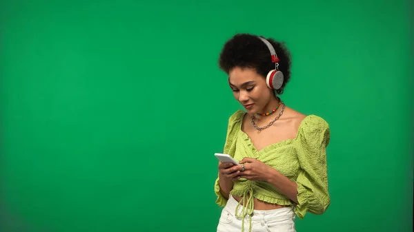 Ricci donna afroamericana in camicetta e cuffie senza fili utilizzando smartphone isolato su verde — Foto stock
