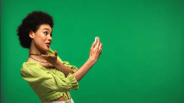 African american woman in blouse looking at smartphone during video chat isolated on green — Stockfoto