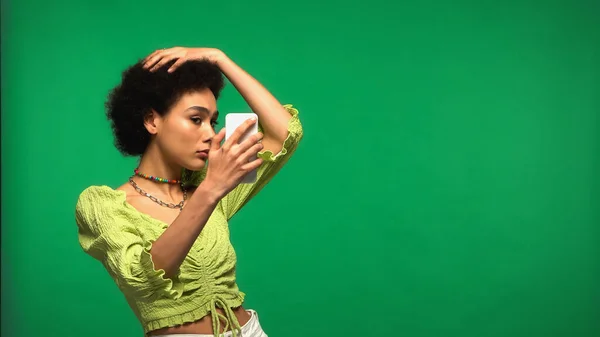 Young african american woman in blouse looking at smartphone isolated on green — стокове фото