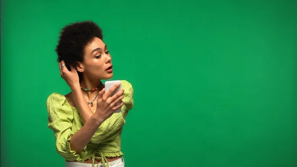 African american woman in blouse looking at smartphone and adjusting hair isolated on green — стокове фото