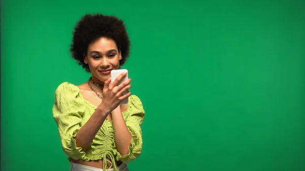 Mujer afroamericana complacida en blusa mirando teléfono inteligente aislado en verde - foto de stock