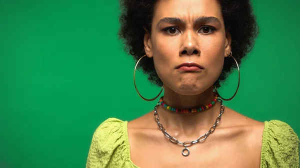 Femme afro-américaine en colère dans des boucles d'oreilles cerceau regardant la caméra isolée sur vert — Photo de stock