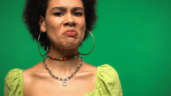 Femme afro-américaine mécontente en boucles d'oreilles cerceau regardant la caméra isolée sur vert — Photo de stock