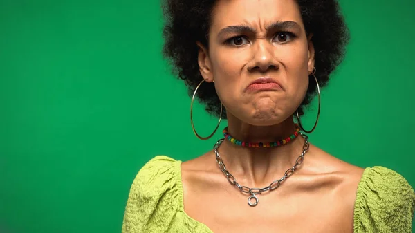 Femme afro-américaine en colère dans des boucles d'oreilles cerceau grimacing isolé sur vert — Photo de stock