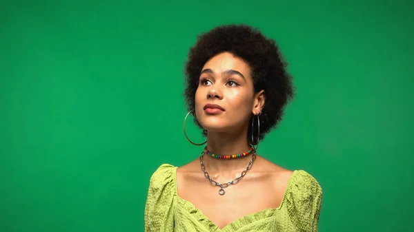 Sognante donna afro-americana in orecchini a cerchio guardando altrove isolato sul verde — Foto stock