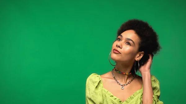 Mujer afroamericana soñadora en pendientes de aro ajustando el pelo rizado aislado en verde - foto de stock