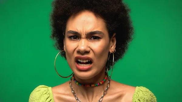 Upset african american woman in hoop earrings looking at camera while grimacing isolated on green — Foto stock