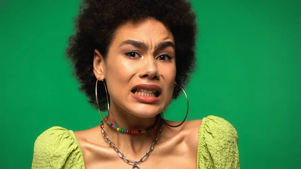 Незадоволена молода афроамериканська жінка в сережках обруча, дивлячись на камеру ізольовано на зеленому — стокове фото