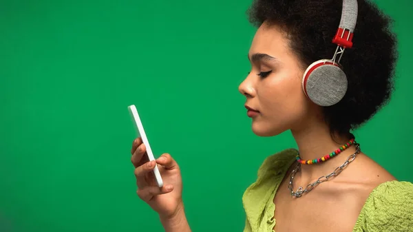 Vista laterale della donna afro-americana in cuffie wireless ascoltare musica e guardare smartphone isolato su verde — Foto stock