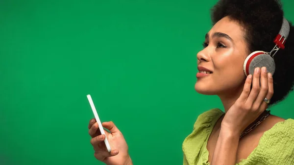 Pleased african american woman in wireless headphones listening music and looking at smartphone isolated on green — Stock Photo