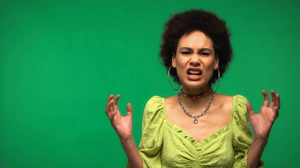 Mujer afroamericana irritada en pendientes de aro gestos y mirando a la cámara aislada en verde - foto de stock