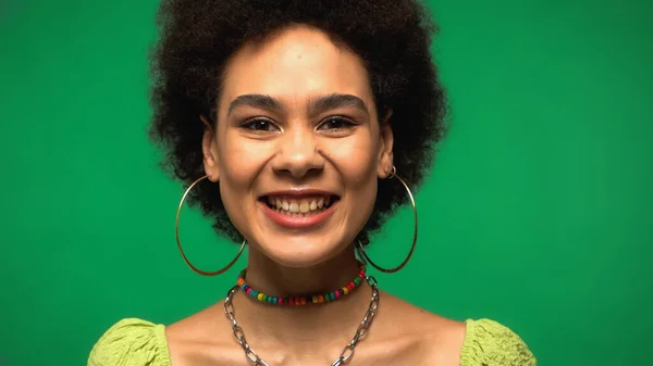 Mujer afroamericana positiva en pendientes de aro mirando a la cámara aislada en verde - foto de stock