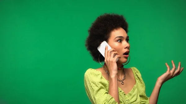 Shocked african american woman in hoop earrings talking on cellphone isolated on green — Stockfoto