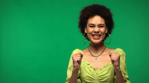 Excited african american woman in hoop earrings smiling isolated on green — стоковое фото