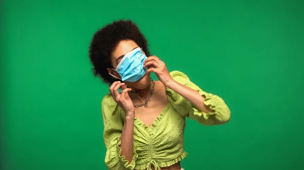 Curly african american woman wearing blue medical mask isolated on green — Fotografia de Stock