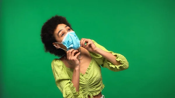 Jovem afro-americana vestindo máscara médica azul isolado no verde — Fotografia de Stock