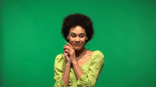 Sonriente mujer afroamericana en blusa y pendientes de aro mirando hacia otro lado aislado en verde - foto de stock