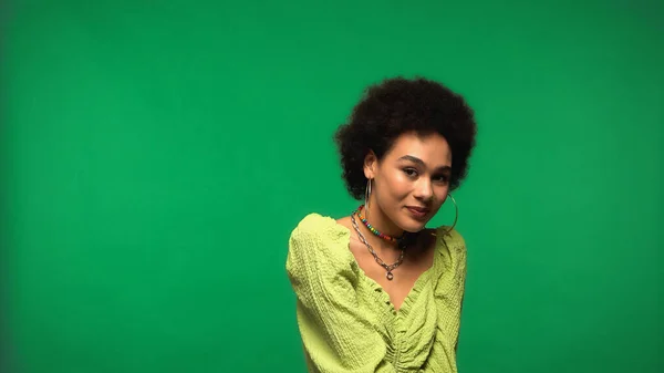 Positive african american woman in blouse and hoop earrings smiling isolated on green — Stockfoto