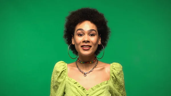 Happy young african american woman in blouse and hoop earrings smiling isolated on green — Stockfoto