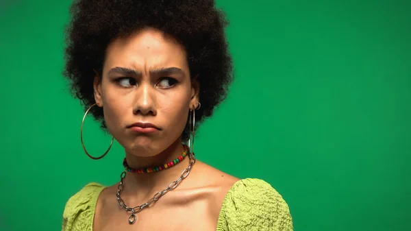 Mujer afroamericana disgustada en blusa y pendientes de aro mirando hacia otro lado aislado en verde - foto de stock