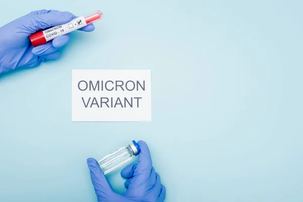 Top view of doctor in latex gloves holding test tube and vaccine vial near card with omicron variant lettering on blue — Photo de stock