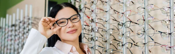 Happy asian oculist looking at camera and touching eyeglasses in optics salon, banner — Stock Photo