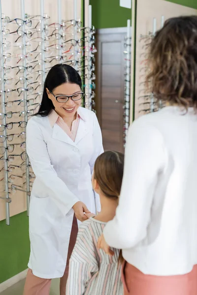 Happy asian oculist in eyeglasses holding hand of girl near blurred mother in optics store — Stock Photo