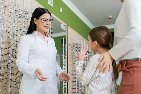 Smiling asian oculist talking to girl and mother near assortment of eyeglasses in optics store — Stockfoto