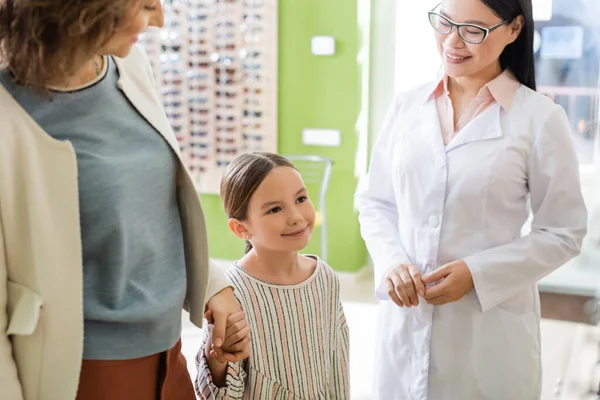 Pleased mother and daughter holding hands near asian oculist in optics store — Stock Photo