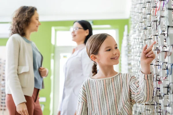 Smiling girl pointing at assortment of eyeglasses near blurred mom and asian oculist in optics store — Stockfoto